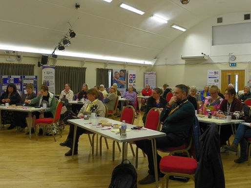 Audience attentively listening in a conference room with red chairs. NHS banner in background. People taking notes, focused and engaged.
