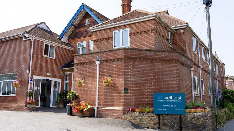 Photograph of a red brick nursing and residential home named Ivelhurst, featuring a two-storey building with white-framed windows and a chimney. A blue sign with white text is positioned near the entrance, alongside flower pots and hanging baskets adding colour to the exterior.
