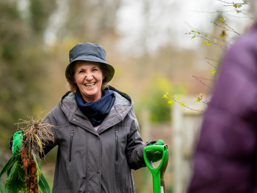 Smiling woman in a hat and grey coat holds carrots and a green shovel outdoors. Blurred natural background and another person visible.