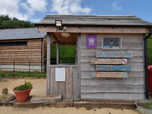Photograph of a small wooden entrance booth with signs reading "WELCOME TO ARK" and "VISITS BY APPOINTMENT" under a partly cloudy sky. Surrounding elements include potted plants, a stone sculpture, red bins, and a wooden fence, indicating a rustic outdoor setting.