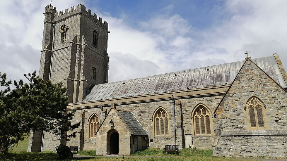 Saint Andrews Church Burnham on Sea. A historic stone church featuring a tall square tower with battlements and a clock, alongside a pitched roof and arched windows. The scene includes a grassy foreground with bushes and a tree under a partly cloudy sky.