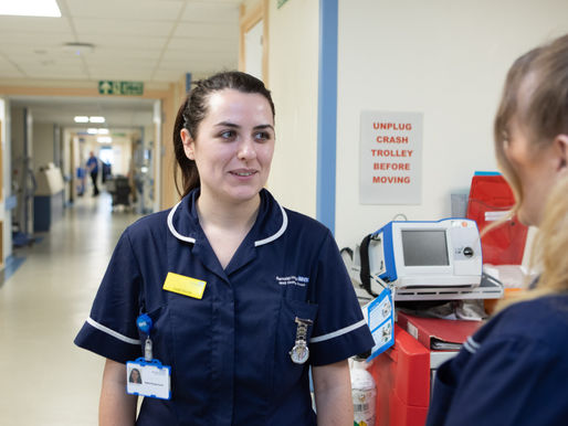 Two nurses in a hospital corridor chatting. One smiles, wearing a navy uniform. A sign reads "UNPLUG CRASH TROLLEY BEFORE MOVING."