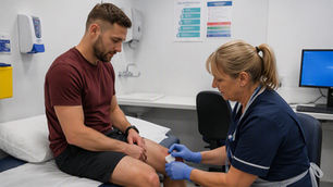Nurse in blue gloves cleans a man's knee wound in a clinic exam room while he sits on the bed.