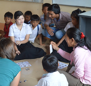 Girl with down syndrome central in focus seated on the floor surrounded by other children, a female teacher holding a flashcard and two other adults look on