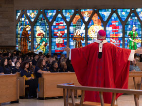 A priest in red garments with arms wide in homily to an audience. Stained glass windows in the background.