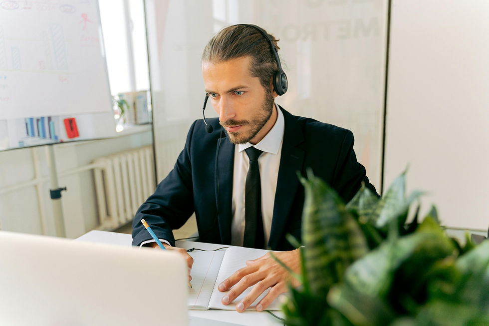 An assistant with headset sits at desk writing in notebook, focused.
