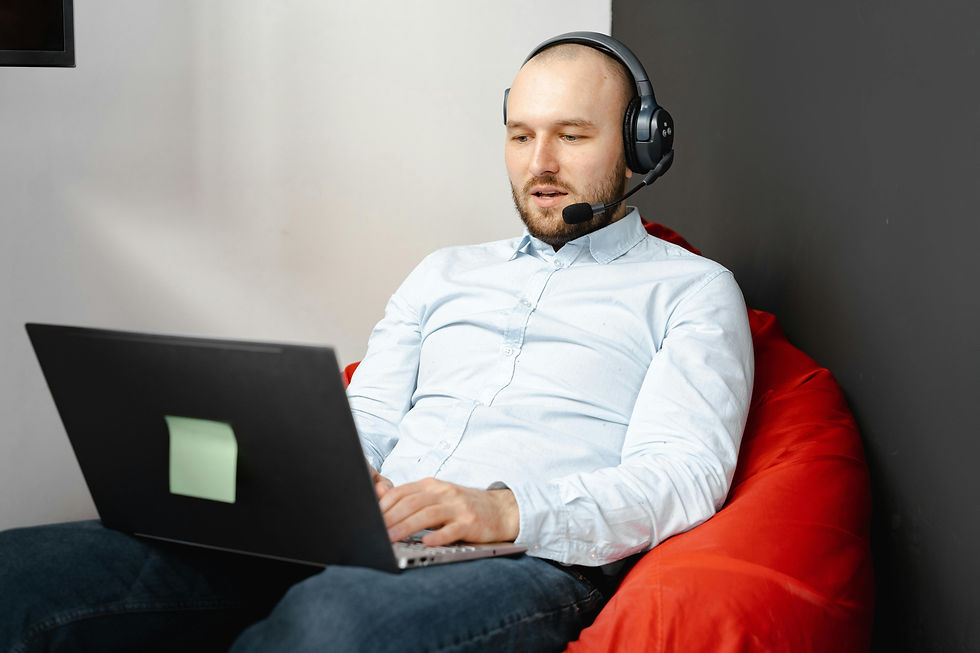An assistant sits on a red bean bag, wearing a headset, typing on a laptop.