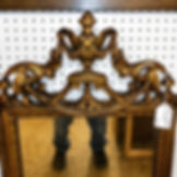 Close-up of the top crest on a gold gilt wall mirror with carved urn detail, scrollwork, floral accents, and aged gilt finish