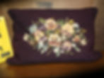 View of a needlepoint pillow with light colored flowers and dark material.