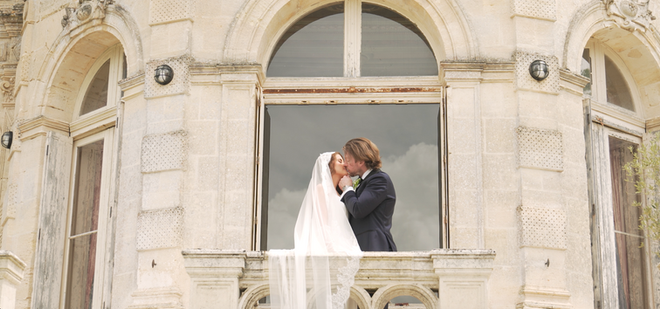 Bride and groom kissing on the balcony of Château de la Valouze with the bride’s veil flowing in the wind.
