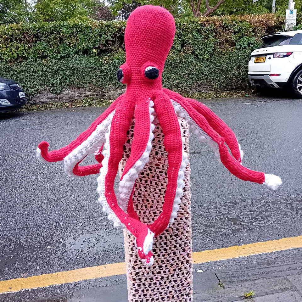 A red crochet octopus atop a bollard  covered with pink crochet. with a road behind