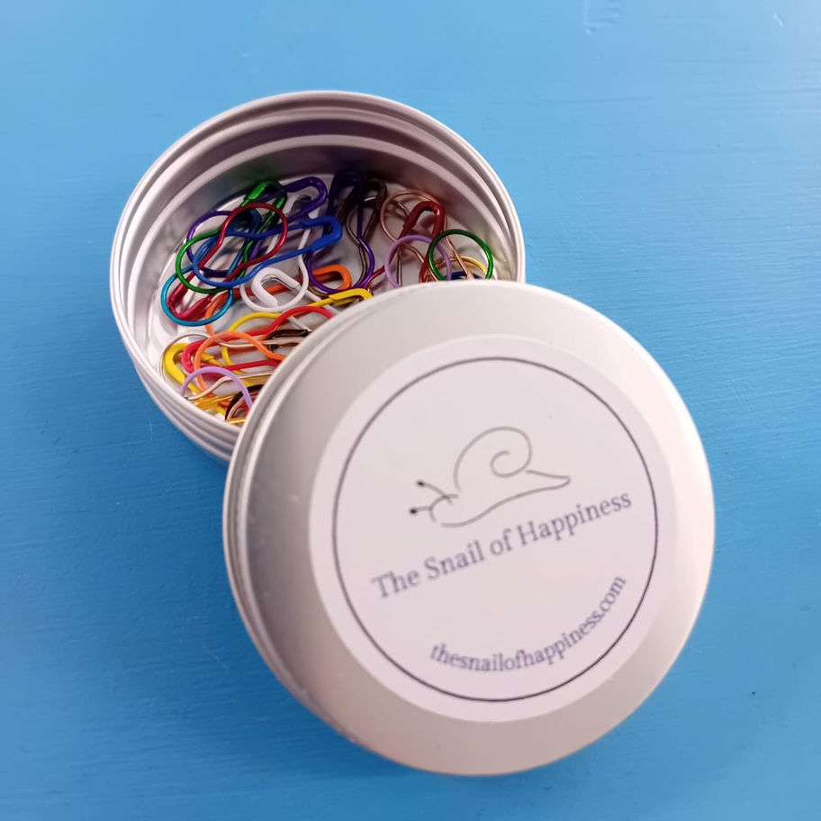 Open Snail of Happiness round tin, with the lid partially covering the contents, with coloured metal stitch markers showing