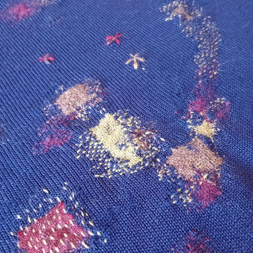 Blue knitted jumper repaired with darning and hand-stitched embroidery features red, yellow, and beige patches in various shapes, creating a decorative pattern.
