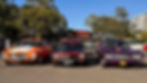 Three vintage cars, orange, black, and purple, are parked side by side in a sunlit lot with trees and buildings in the background.
