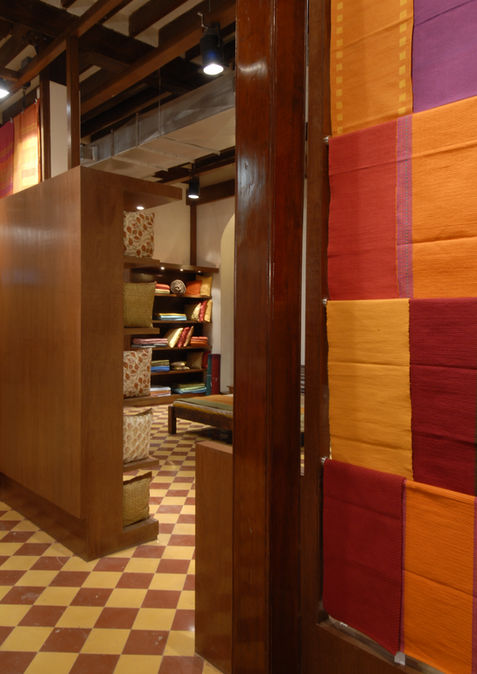 Checkerboard tile flooring, exposed wooden beams, plaster walls, warm wood partitions, wooden bench with cushions, recessed textile displays for apparel store, Linear recessed downlights, adaptive re-use retail store, retail store inspired by indian context, indian home decor store fro bedroom, Woven fabrics suspended in retail store, baffle ceiling with exposed services in retail store