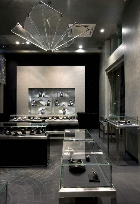 Double height retail space, monochromatic material palette in retail space, textured subtle walls, mezzanine floor in retail store, dark stone flooring in jewellery store, branded motifs in diamond store, minimalist sleek furnishing in retail store, suspended glass pendants chandeliers, glass cases displaying diamond pieces, cobblestone inspired flooring tile patterns in retail store