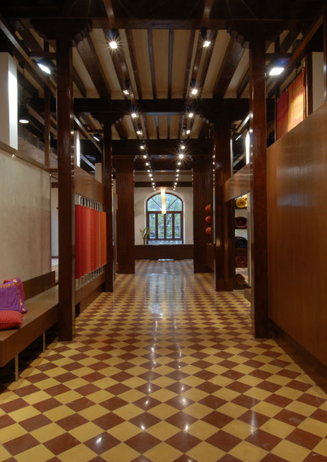 Checkerboard tile flooring, exposed wooden beams, plaster walls, warm wood partitions, wooden bench with cushions, recessed textile displays for apparel store, circular downlights, natural light from arched window, adaptive re-use retail store, retail store inspired by indian context
