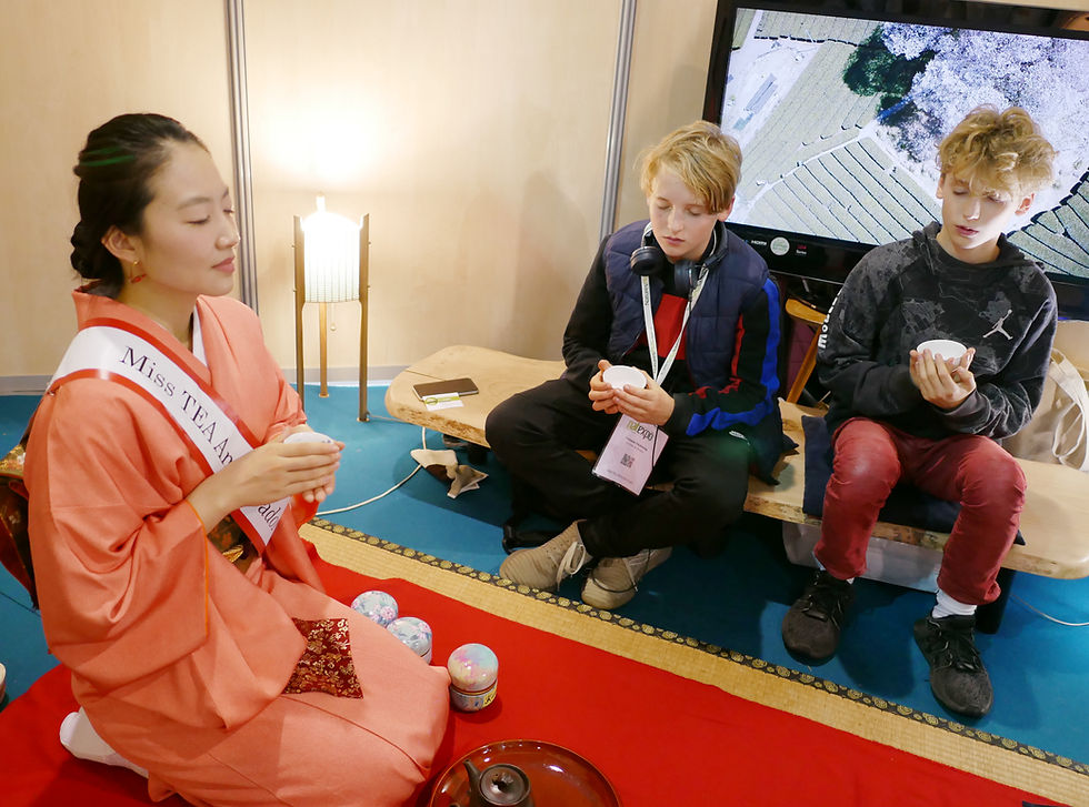 A woman in a kimono and two boys sit on mats holding cups, participating in a tea ceremony. A sign reads "Miss TEA Ambassador."