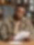 Man in camo jacket reading documents at a wooden table, kitchen background. Appears thoughtful, focusing on papers in hand.