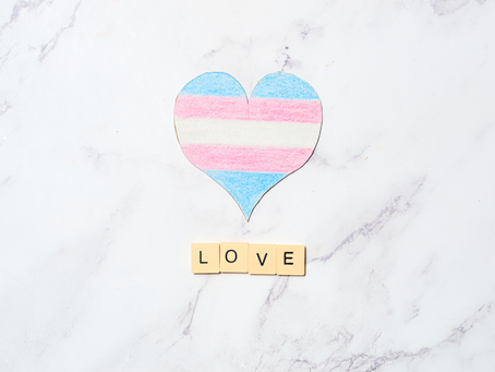 Heart with transgender flag colors on marble background. Below, Scrabble tiles spell "LOVE." Emotive and supportive theme.