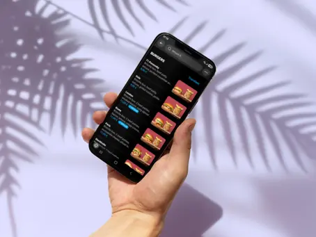 Hand holding a smartphone displaying burger menu with prices. Background has leaf shadows on a light surface, creating a relaxed vibe.