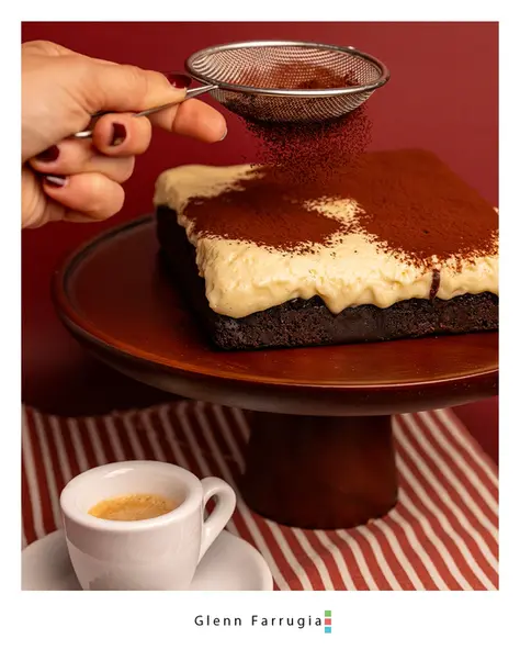 Hand dusting cocoa powder over a tiramisu brownie topped with cream, served alongside a cup of espresso.