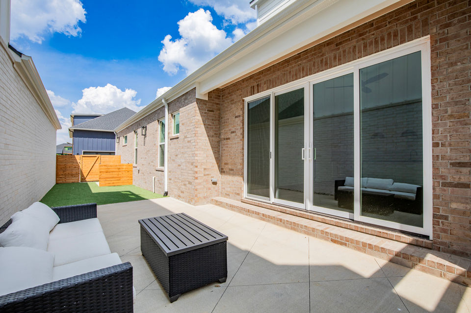 Outdoor courtyard with a sectional sofa and coffee table facing sliding glass doors, with turf lawn and wood fencing in the background.