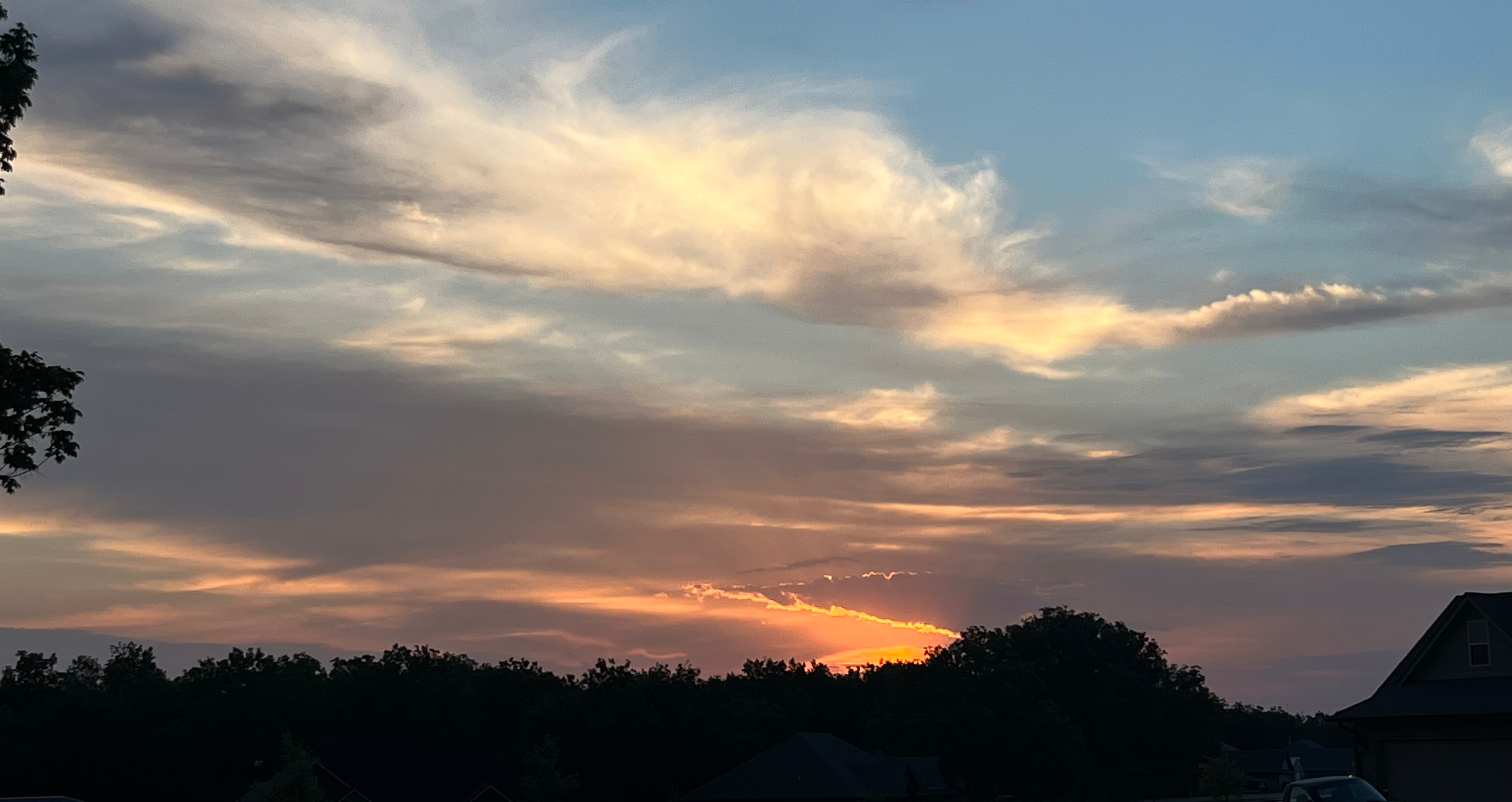Sunset over a tree-lined neighborhood with warm orange light near the horizon and soft blue, gray, and golden clouds stretching across the sky.
