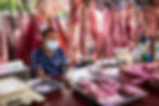 Un vendedor de carne en un mercado local, rodeado de cortes de carne cruda colgando y en bandejas. La imagen destaca la conexión entre los hábitos alimentarios y las enfermedades zoonóticas, como se discutió ampliamente durante la pandemia de COVID-19.