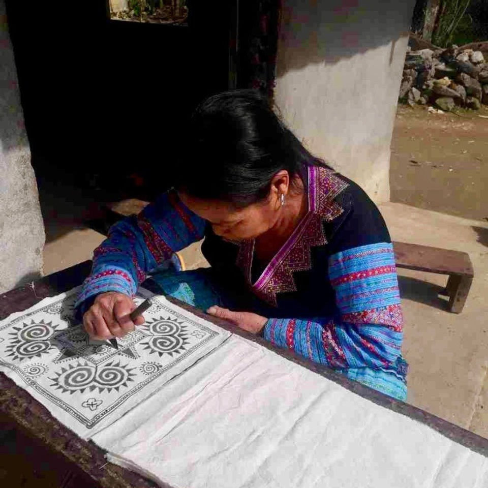 A photograph of a lady from the Hmong People of Hao Binh, stencilling a cushion cover with a traditional story of the elements Earth, Sky, Work and family.