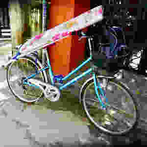 A photograph of a blue bike with an ironing board secured on with bungee elastic.