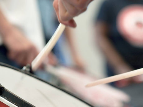 Close-up of hands playing a red-and-black drum with drumsticks, blurred people in the background.