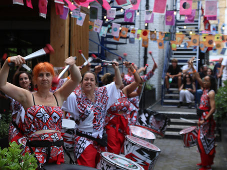 Batala London drummers in red-and-white outfits drum and raised sticks in a decorated alley with colourful bunting and lights.