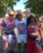 Four smiling carnival-goers in red, black and white Batala London outfits pose on a sunny street under trees.