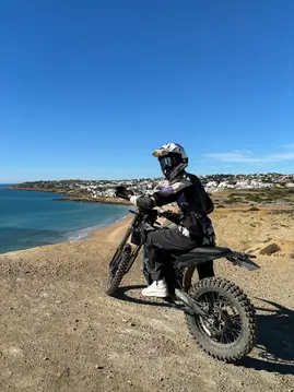 a motocross rider in helmet looking at the camera on a cliff with blue sea, a town of white buildings and sky behind