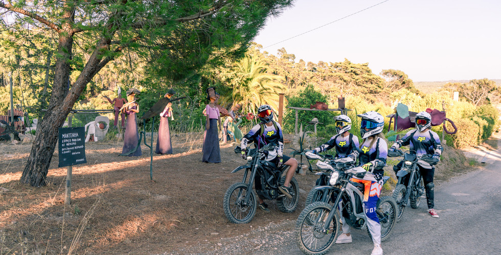 three persons in motocross gear on the bikes surrounded by trees and large sculptures of humans and animals
