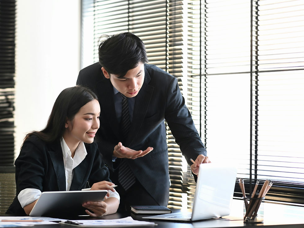Two professionals in suits collaborate at a desk. One holds a tablet, the other gestures towards a laptop. Bright setting with blinds.