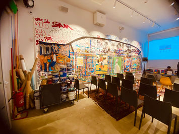 Foto della sala eventi con sedie vuote. Una parete è interamente decorata con dipinti di vario genere.