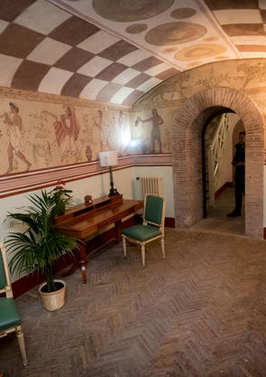 Foto di uno dei locali di Palazzo Migliori: si vedono pareti e soffitto affrescati, una porta ad arco, una scrivania e due sedie sul lato lungo.