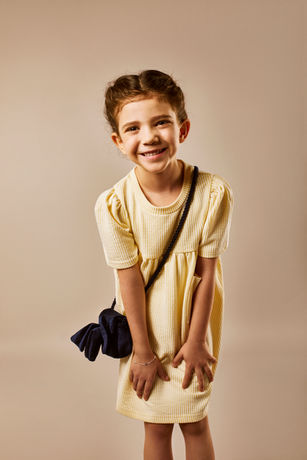 a little girl wearing a yellow dress and a blue purse smiles MAF