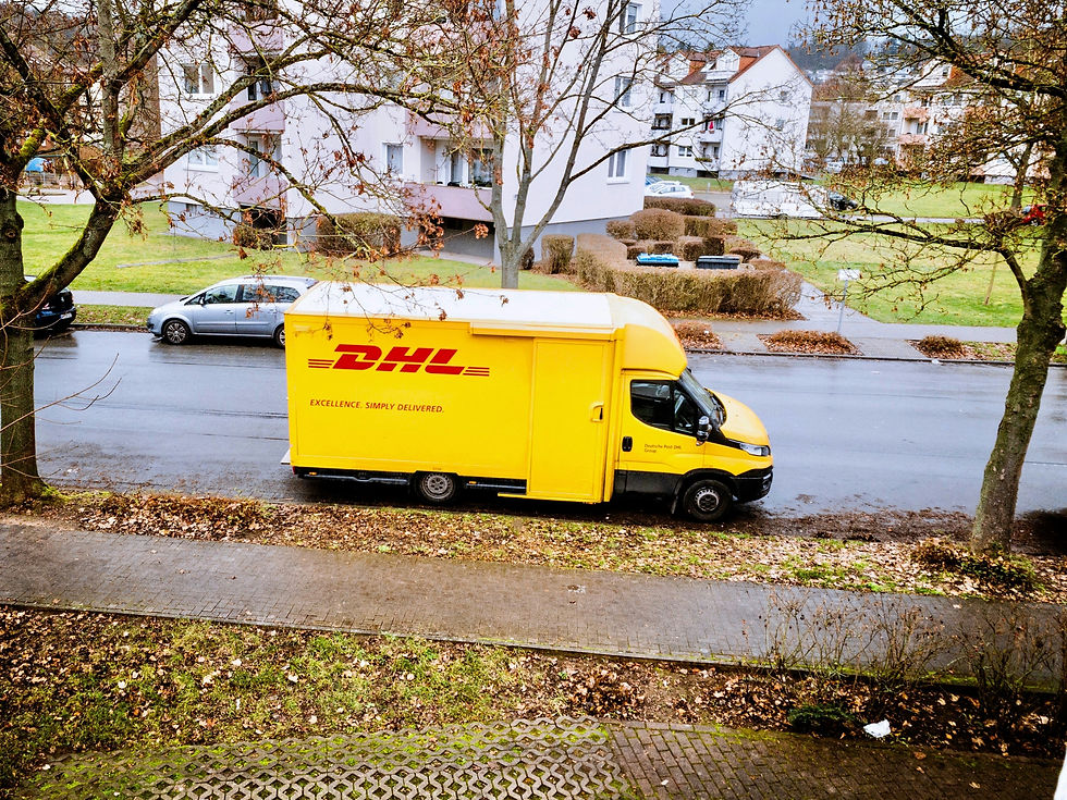 Yellow DHL truck parked on a street with bare trees and grass. Buildings in the background. Text: "Excellence. Simply Delivered."