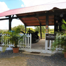 Located on the edge of the N9 near the beaches of the west coast between Grand Bourg and Saint Louis, the small artisanal distillery POISSON with barely 300,000 liters of rum produced per year, is nevertheless a must in Marie-Galante which will take you on a surprising return at the sources. Welcomed by smiling staff, in a green park with old mango trees and colorful flamboyant trees, you will immediately be won over by the picturesque and friendly atmosphere that emanates from the place. During a completely independent visit, like a treasure hunt, you will gradually discover the memory of this estate and the circuit of a rum with the flavors of yesteryear.
Sugar factory founded in 1860 by the Poisson family, the estate was transformed into a distillery in 1916 when it was bought by Edouard Rameau, descendant of 18th century settlers. The latter added a steam engine and a copper still and baptized his Rum under the name well known to initiates of “Père Labat”, in memory of a French missionary installed on the island at the end of the 17th century.
The few renovations made by the successors of E. Rameau will in no way distort this cultural heritage and it is at the heart of this history of more than a century that you will be immersed. Here, the witnesses of the past have in fact remained the same: the still and the recently stopped steam engine are still in place. The sugar boilers are still in their massive masonry and the thick-walled buildings as well as the main wooden house bear witness to their age.