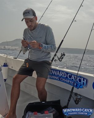Un cliente concentrado revisando el hilo de su caña de pescar mientras está sentado cómodamente en la banda de babor de Windward Catch Fishing Charter con las costa de Tenerife de fondo.
