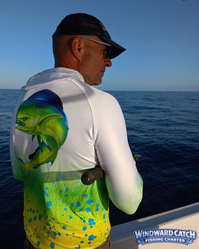 The captain in profile wearing a cap and white sweatshirt with a drawing of a goldfish, holding a fishing rod from the side of the ship in the open sea.