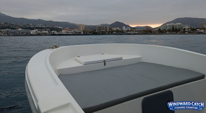 Detalle de las colchonetas grises acolchadas y tambucho de proa del barco con el mar y la costa de fondo al amanecer.