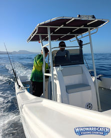Vista lateral de la cubierta del barco blanco de Windward Catch en navegación con el capitán en el puesto de mando y un cliente al lado, mostrando cañas de pescar y la costa al fondo.