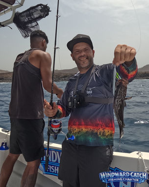 Cliente con su propia marca local Vulcan Lures de señuelos en Canarias, con gorra negra y camiseta técnica multicolor sonríe mientras sostiene un pez serrano en la popa del barco Windward Catch Fishing Charter, con otro compañero de espaldas detrás.
