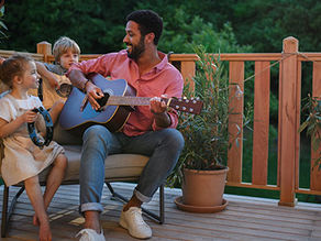 Young African american man enjoys playing the guitar with little children accompaining him with instruments on patio in garden in summer.