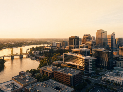 Golden-hour aerial of a downtown skyline beside a river and bridge, with glass towers glowing under a hazy sky.