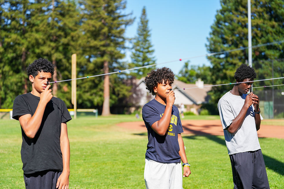 Three young men stand on a sunny field, holding strings to their noses in a focus exercise, with trees and a baseball diamond behind them.